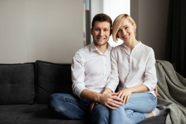 Eşleşen beyaz gömlekler ve kot pantolon giyen mutlu bir çift modern bir koltukta yakın oturuyorlar, birlikte vakit geçiriyorlar ve sıcak bir yuva kuruyorlar..