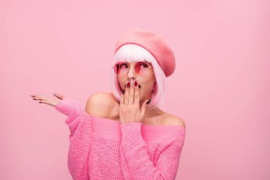 Canlı pembe bir elbise giymiş, gözlüklü ve şapkalı şık bir kadın, benzersiz ve modern tarzını sergileyerek, uyumlu, pembe bir arka plana karşı anlamlı bir şekilde poz veriyor..