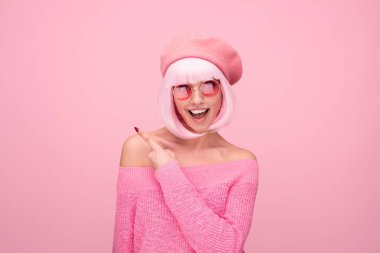 Pembe elbiseli ve aksesuarlı neşeli bir kadın pembe bir arka plana karşı heyecanla işaret ediyor. Pozitif ve neşe gösterir.