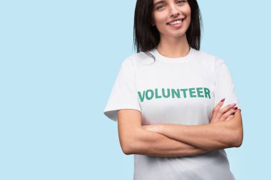 Üzerinde gönüllü olan bir tişört giyen genç bir kadın özgüvenle gülümsüyor ve açık mavi bir arka plana karşı poz veriyor, yardım, bağış ve toplumsal katılımın temaları ifade ediliyor.
