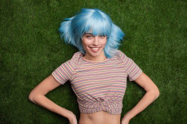 Canlı, mavi saçlı genç bir kadın, yemyeşil çimenlerin üzerinde uzanmış, neşeli çizgili bir bluz giyiyor, neşe saçıyor ve bireysellik yayıyor..