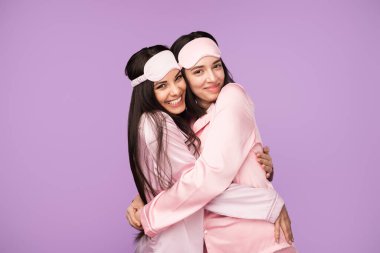 Pembe uyku giysisi giyen iki neşeli kadın açık mor bir arka planda sarılıp gülümsüyor. Aynı pijama takımları ve uyku maskeleri sıcak ve neşeli bir dostluk atmosferi yaratıyor.