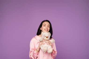 Pembe pijama giyen neşeli genç bir kadın bir oyuncak ayıyı kucağında tutuyor. Mor bir arka plana sıcak bir şekilde gülümsüyor. Rahatlık, neşe ve nostalji hissi uyandırıyor..