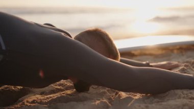 Dalgıç kıyafeti giyen bir sörfçü sörf seansından sonra gün batımında plajda gerilir. Sörf yaptıktan sonra plajda yoga yapıyor..