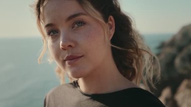 Genç bir kadının başını kameraya çevirdiği, objektife sakin bir ifadeyle baktığı yakın plan. Okyanus ve uçurum arkaplanı.