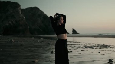 Gün batımında bir dansçı zarif bir şekilde sahilde hareket eder. Arka planda okyanus ve kayalık uçurumlar, sakin ve sanatsal bir sahne yaratır..