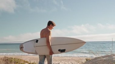 Genç adam sörf tahtasıyla sahilde yürüyor, dalgaları yakalamaya hazır. Bu video yazın özünü, sörfü ve okyanus yaşamının neşesini yakalar..
