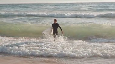 Dalgıç kıyafeti giyen bir sörfçü okyanustan dalgaların altında çıkar. Kıyıya doğru giderken sörf tahtasını kolunun altında taşıyor..