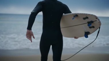 Dalgıç kıyafeti giyen bir sörfçü sörf yapmak için okyanusa koşar. Sörf tahtasını kolunun altında taşıyor. Deniz dalgalı ve gökyüzü bulutlu..