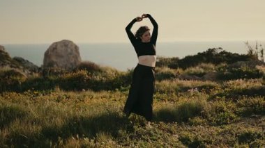 Bir kadın gün batımında bir tarlada zarif bir şekilde dans ediyor, manzaralı kayalık bir manzara ve arka planda okyanusla..