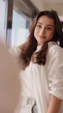 Siyah saçlı neşeli bir kadın aynadaki yansımasına bakarken saç stilini ayarlar ve özgüveniyle hazırlanır..