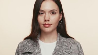 İnce bir gülümsemesi olan güzel, genç bir kadın. Gözlerini açıyor ve kameraya bakıyor. Tarafsız bir bej arka planda kendine güven ve pozitiflik yayıyor..