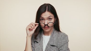 Gözlüklerinin üzerinden süzülen sorgulama bakışlı şık bir genç kadın. Tarafsız bej arka plan modern, entellektüel havayı geliştirir..