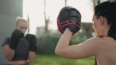 İki kadın dışarıda boks antrenmanı yapıyor, eldiven takıyor ve güneşin altında antrenman pedleri kullanıyor..