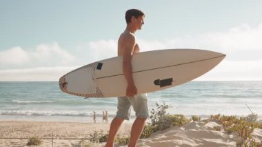 Kum tepeciğinde durup okyanus dalgalarını izleyen bir sörfçü. Sörf tahtasını tutuyor ve gülümsüyor. Sahilde arka planda başkaları da var..