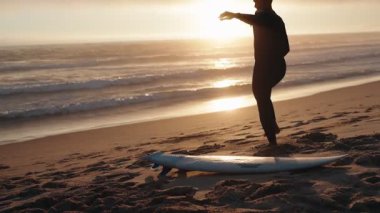 Gün batımında sahilde ısınan bir sörfçü okyanusa girmeye ve dalgalara binmeye hazırlanıyor. Sörf, spor ve seyahat videoları için mükemmel..