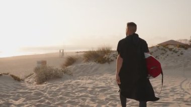 Bir sörfçü gün batımında kum tepelerini aşarak okyanusa doğru yürür, sörf tahtasını taşır. Siyah kapüşonlu panço havlusu giyiyor..