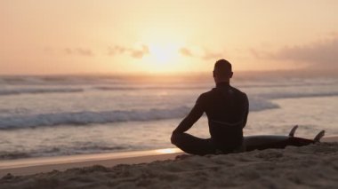 Gün doğumunda sörf tahtasıyla sahilde oturan bir sörfçü dalgalara çarpmadan önce huzur ve sükunet bulur..