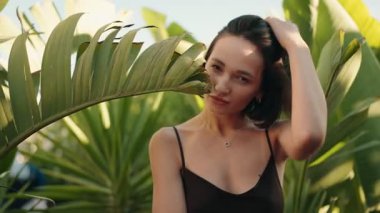 Siyah elbiseli güzel esmer kadın güneş ışığında tropikal bitkilerin arasında poz veriyor, baştan çıkarıcı bir şekilde kameraya bakıyor.