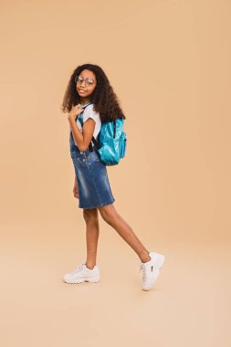 Kendine güvenen genç bir kız elinde mavi bir sırt çantasıyla, şık gözlüklerle, tarafsız bir arka plana doğru gülümsüyor, gündelik moda ve gençlik enerjisini vurguluyor..