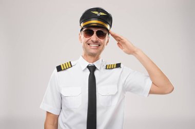 Üniformalı ve güneş gözlüklü profesyonel bir ticari pilot selam verirken, havacılık, adanmışlık ve hizmeti temsil ederken özgüvenle gülümsüyor. Hava yolculuğu ve profesyonellik temaları için mükemmel bir görsel tema..