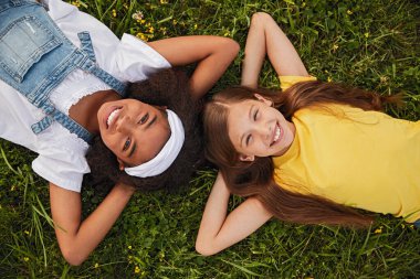 İki neşeli kız yemyeşil çimlerde uzanıyor, gülümsüyor ve açık havada güneşleniyorlar. Barışçıl bir ortamda çekilmiş sembolik bir arkadaşlık ve mutluluk anı..