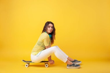 Genç bir kadın, parlak sarı bir zemin önünde kaykayın üzerinde otururken poz veriyor, gündelik ve renkli kıyafetler giyiyor, modern yaşam tarzını, sporu ve dinamik enerjiyi anımsatıyor..