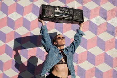 Kot ceketli bir kişi kafasının üstünde renkli bir geometrik duvar taşıyan bir teyp taşıyor..