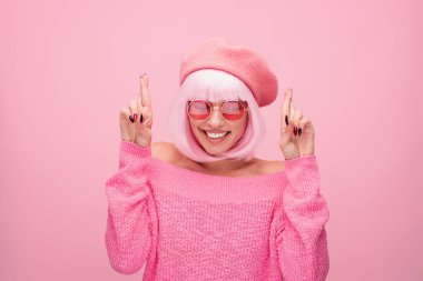 Pembe giyinmiş neşeli genç bir kadın gülümser ve parmaklarını çaprazlar, iyimserlik, eğlence ve modaya uygun cazibeyle dolu neşeli ve neşeli bir sahne yaratır..