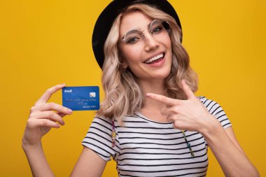 Elinde bir kredi kartı olan neşeli genç bir kadın modern bankacılığı sembolize ediyor. Parlak ve özgüvenli bir şekilde gülümsüyor, cesur ve parlak sarı bir arkaplan tarafından vurgulanıyor..