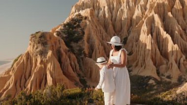 Bir anne ve küçük oğlu güzel kaya oluşumları olan bir sahile bakan bir uçurumda duruyorlar. İkisi de beyaz giyiyorlar ve manzaranın tadını çıkarıyorlar..