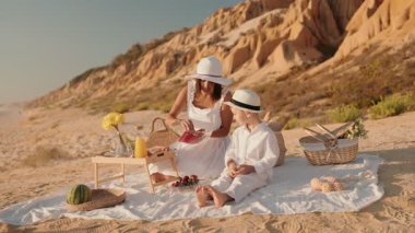Sahilde gün batımında, beyaz elbiseli bir anne ve şapkalı bir anne, beyaz bir sepet, karpuz, çiçekler ve battaniye üzerinde diğer yiyeceklerle piknik yaparken, oğlu için de bir içecek koyar..