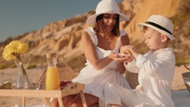 Bir anne ve oğlu bir pastayı paylaşıyor, gülümsüyor ve beyazlar içinde, plaj pikniği sırasında, sevgi, bağ ve yaz eğlencesi duyguları uyandırıyor..