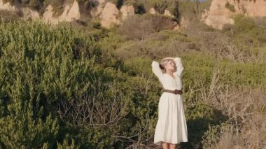 Beyaz elbiseli baştan çıkarıcı sarışın kadın kollarını başının üstüne kaldırmış, doğanın güzelliğini kucaklıyor..