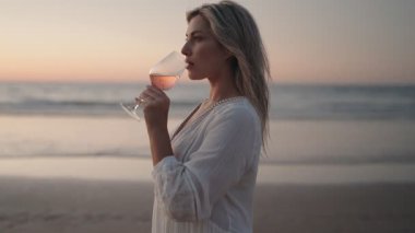 Genç sarışın bir kadın gün batımında sahilde bir bardak gül şarabı içer. Beyaz bir elbise giyiyor ve okyanusa bakıyor..
