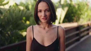 Güzel bir esmer kadın, egzotik bir tropik bahçe ortamında kameraya gülümsüyor. Kendinden emin, mutlu ve tatilinin tadını çıkarıyor..