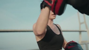 Boks eğitimi alan kararlı bir kadının yakın plan çekimi. Dışarıda antrenör tarafından tutulan boks pedlerine yumruk atıyor..