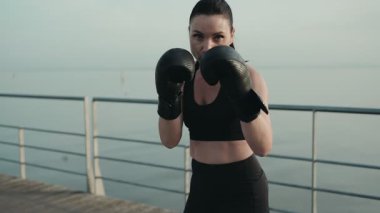 Boks eldivenli, açık havada antrenman yapan, güneşli bir günde denize yumruk atan odaklanmış atletik bir kadın..