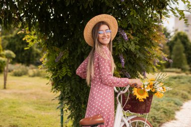 Puantiyeli elbise, güneş şapkası ve gözlüklü neşeli bir kadın çiçek sepeti ile klasik bir bisikletin yanında poz verirken doğal ışık altında serin bir parkın keyfini çıkarıyor..