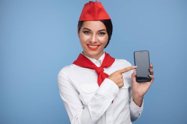 Klasik üniformalı profesyonel bir uçuş görevlisi akıllı telefonu gösterirken gülümsüyor..
