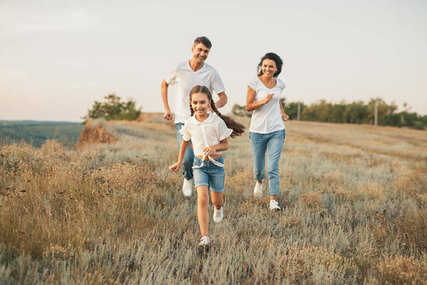Веселая семья проводит время на свежем воздухе, бегает по живописному полю, наполненному зеленью, наслаждается природой и делится улыбками в яркий солнечный день.