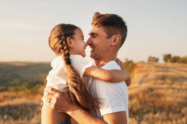 Bir baba ve kızı çarpıcı bir gün batımında açık havada samimi bir kucaklaşmayı paylaşıyor. Sevgi ve mutluluğu huzurlu bir ortamda yakalıyorlar..