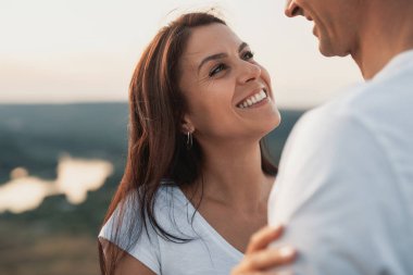 Açık havada romantik bir anı paylaşan mutlu bir çift, arka planda huzurlu bir günbatımı ile kucaklaşıp gülümsüyor..