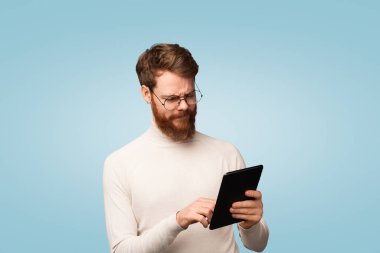 Açık balıkçı yaka giyen sakallı adam, mavi arka planda gösterilen merakını dile getirirken tablet bir cihaz kullanıyor. Teknoloji kullanımı, bilgi arayışı ve modern yaşam tarzını temsil eder.