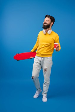 Kaykaylı ve kahve fincanı olan bir adam gülümsüyor, mavi arka planda günlük kıyafetler giyiyor. Yaşam tarzı, kentsel ve boş zaman temaları için ideal.