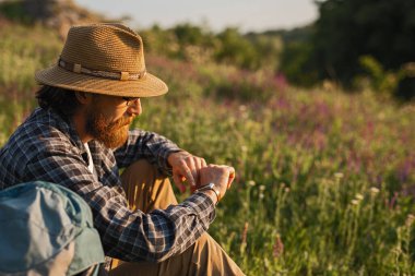 Güneşli bir tarlada oturan sakallı ve gözlüklü bir adam, örülmüş bir şapka ve ekose gömlek giymiş, doğanın güzelliği ile çevrili iken saatine dikkatlice bakıyor..