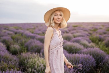 Güneş şapkası takan neşeli genç bir kadın günbatımında elinde narin bir çiçek tutarak lavanta tarlasının tadını çıkarıyor. Işıldayan mutluluk, huzurlu bir ortamda doğanın güzelliğini kucaklıyor..