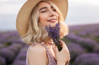 Çiçek açan bir tarlada neşeyle bir demet lavanta çiçeği tutan genç kadın gün batımında doğanın sükunetini ve güzelliğini kucaklıyor..