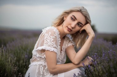 Zarif genç bir kadın çiçek açan lavanta tarlasında oturuyor, narin beyaz dantelli bir elbise giyiyor, güzel doğayla çevrili, huzurlu bir atmosferde güzellik ve stil anları sergiliyor..