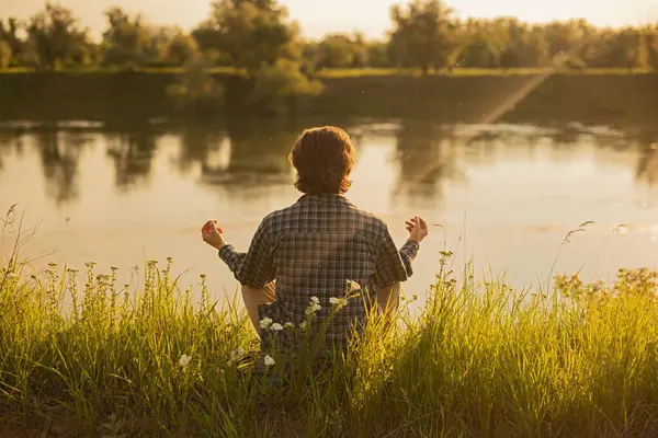 Sakin ve güneşli bir günde çimenli bir nehir kıyısında meditasyon yapan bir insanın huzurlu sahnesi, rahatlamayı ve doğayla olan bağlantısını yansıtıyor..